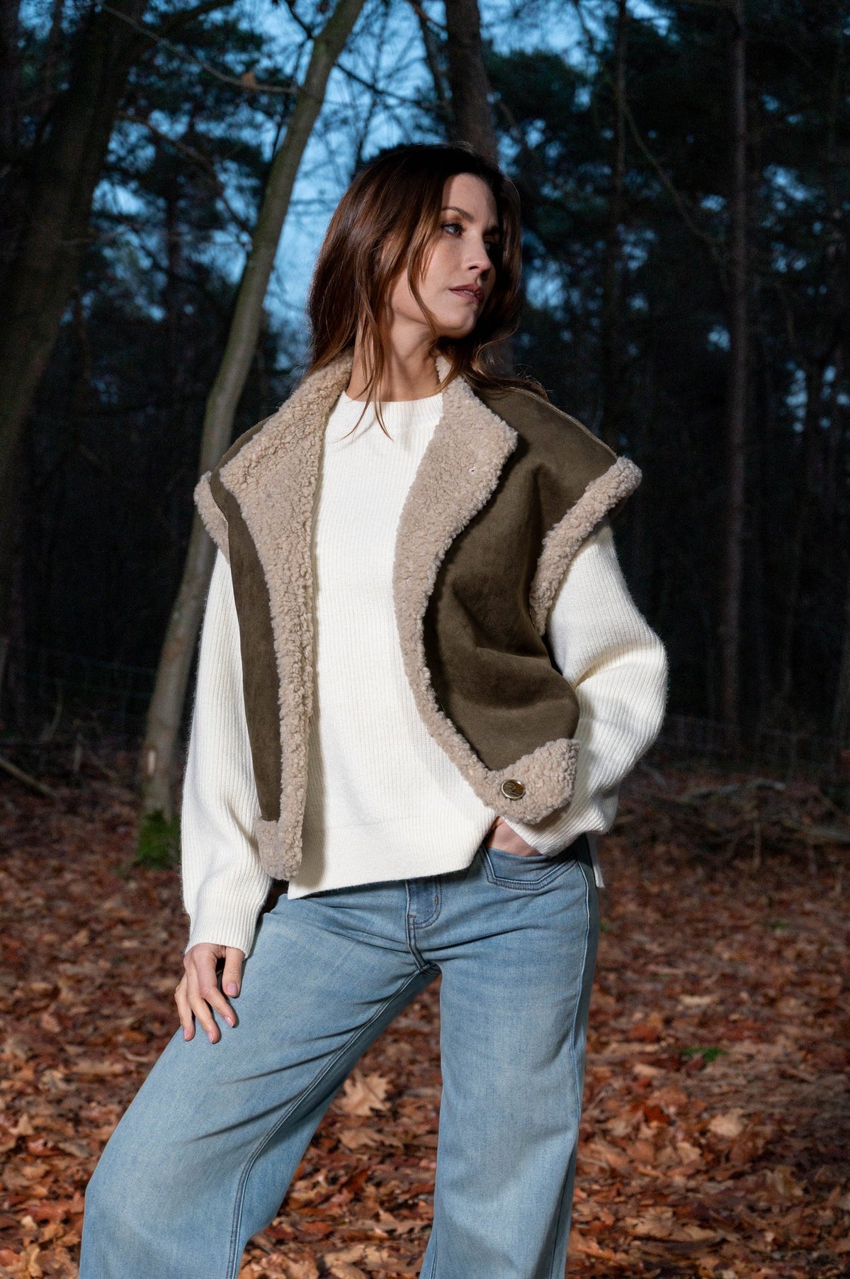 model draagt een mosgroen gilet over een offwhite trui op een lichte spijkerbroek in het bos