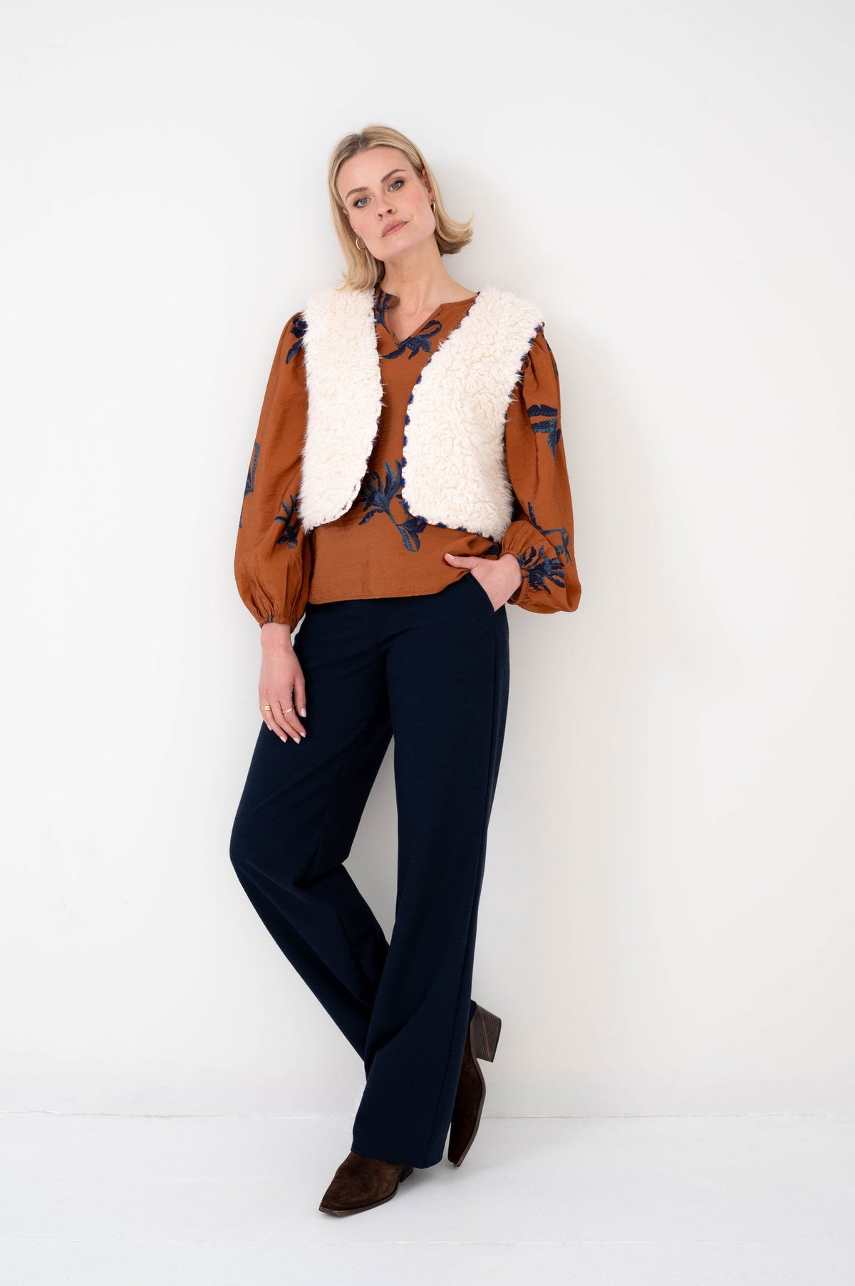dame draagt zedan gilet op een roestbruine blouse met bloemenprint en met een donkere spijkerbroek