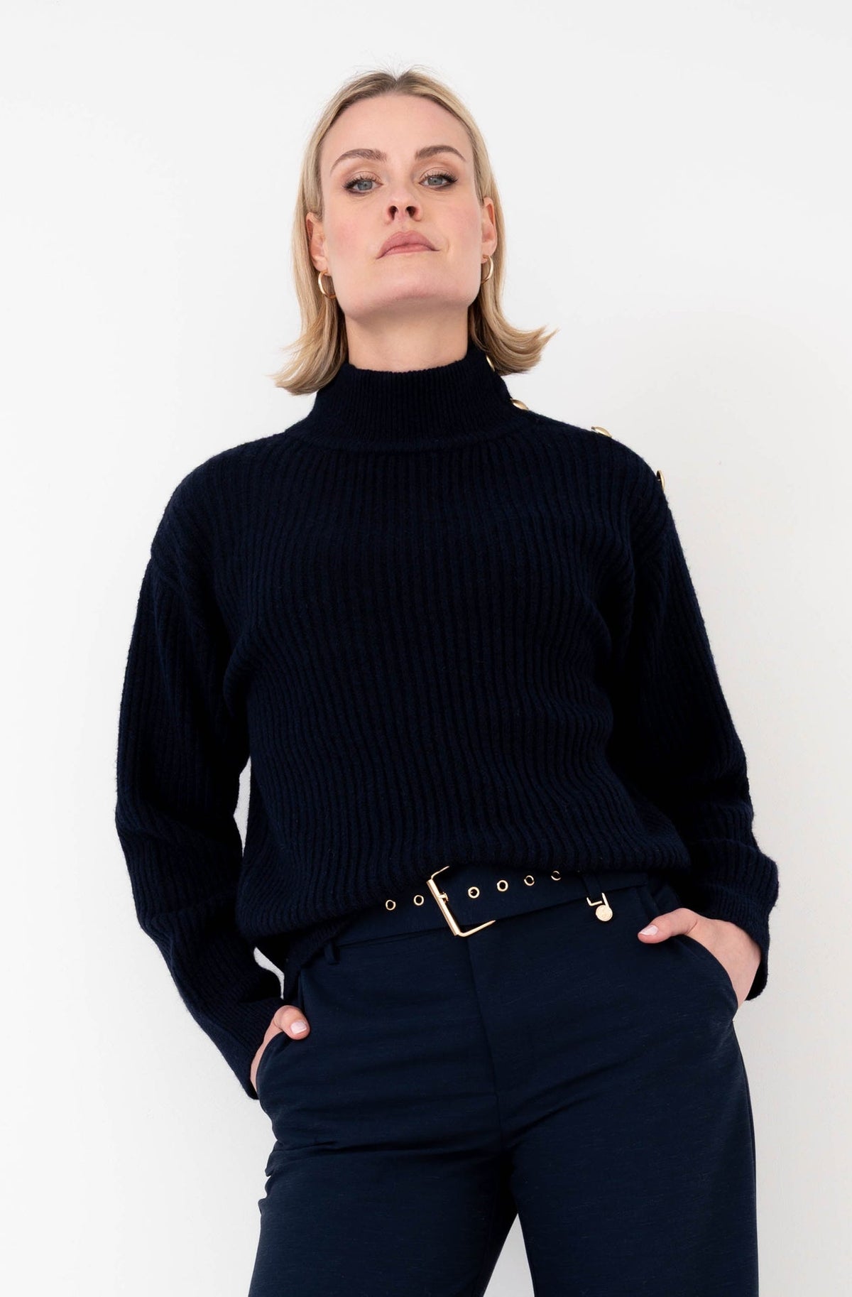 donkerblauwe outfit met een coltrui met ribstructuur en knopen op een schouderkant gecombineerd op een blauwe broek met bijpassende riem